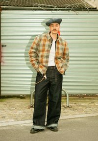 Veste à carreaux vert, orange et beige, associée à un pantalon large en denim foncé. Chaîne d'accent avec un pendentif, et un béret bleu.