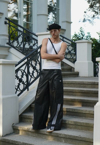 Persona vestida con una camiseta sin mangas blanca y pantalones cargo de cuero negros con tres rayas blancas, de pie en unos escalones de piedra cerca de una barandilla.