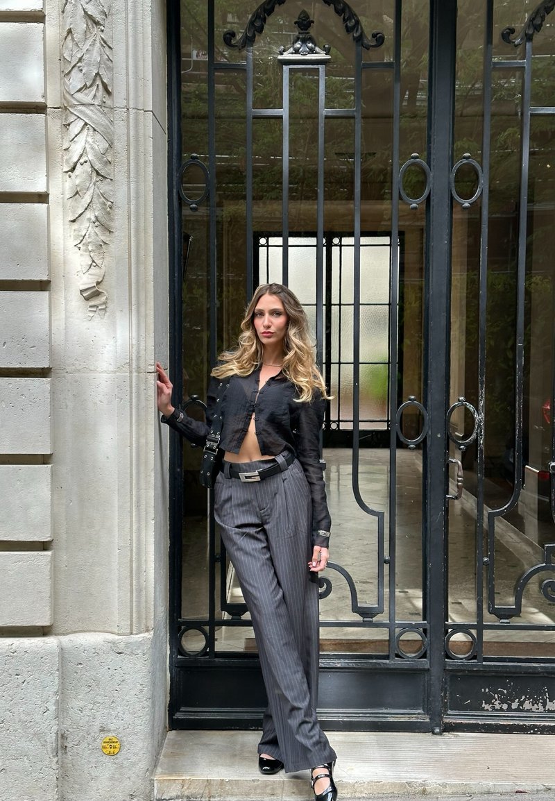 Νέα γυναίκα με μακριά ξανθά μαλλιά, φορώντας μαύρο κοντό μπουφάν και γκρι παντελόνι με ριγέ, ακουμπά σε διακοσμητική μαύρη μεταλλική πόρτα.