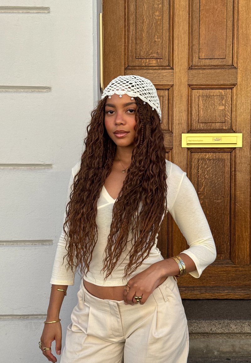 Vrouw met lang krullend haar die een witte gebreide hoofddoek, crème top en broek draagt, staat voor een houten deur met een gouden brievenbusgleuf.