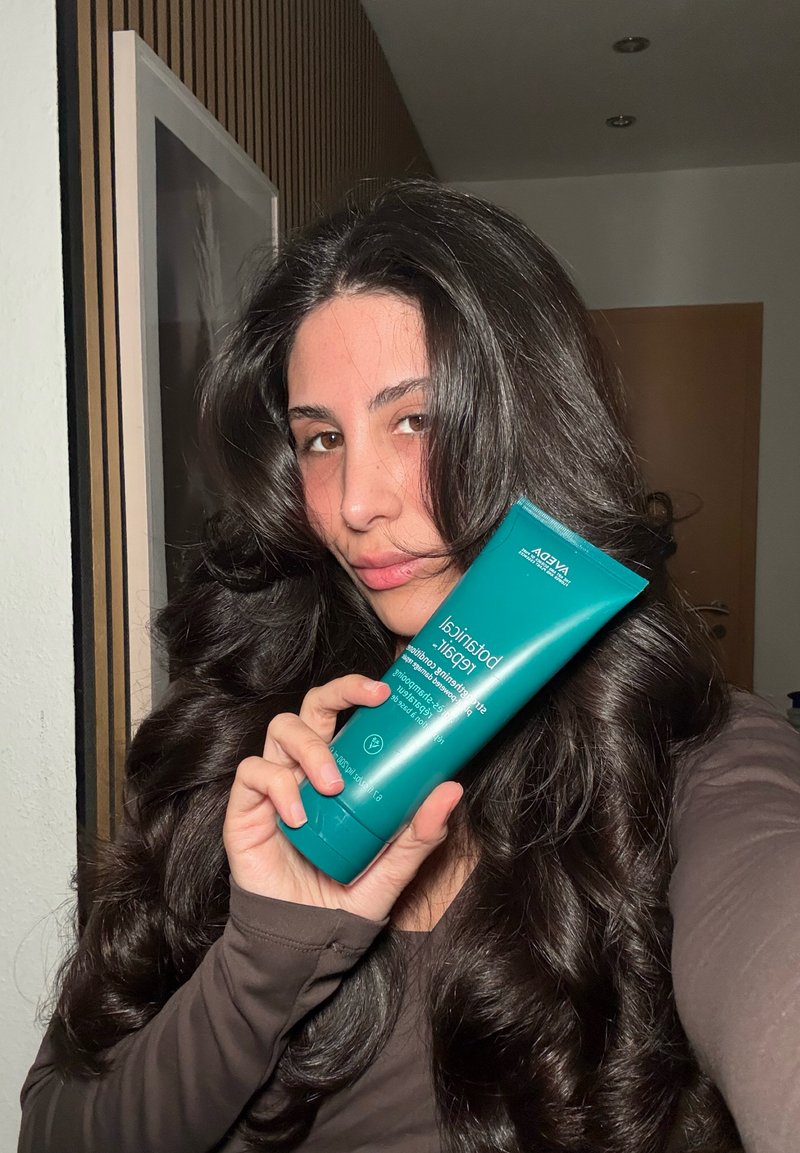 Femme aux longs cheveux foncés tenant un tube teal de soin capillaire Aveda Botanical Repair à l'intérieur, vêtue d'un haut marron à manches longues, debout près d'un panneau mural en bois.