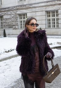 Femme portant un manteau en fausse fourrure violet foncé, des gants en cuir, des lunettes de soleil, tenant un sac à main marron, debout sur un trottoir enneigé près d'un bâtiment en pierre.