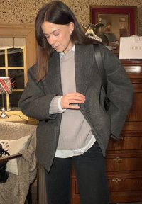 Blazer oversize en laine grise sur un pull gris clair, superposé avec une chemise blanche boutonnée, associé à un pantalon noir. Sac noir à bandoulière à la main.