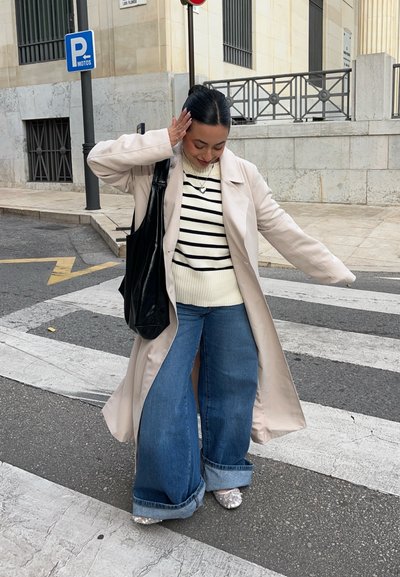 Trench beige claro sobre un suéter blanco a rayas, combinado con unos vaqueros de pierna ancha y zapatos metálicos brillantes. Bolsa tote negra.