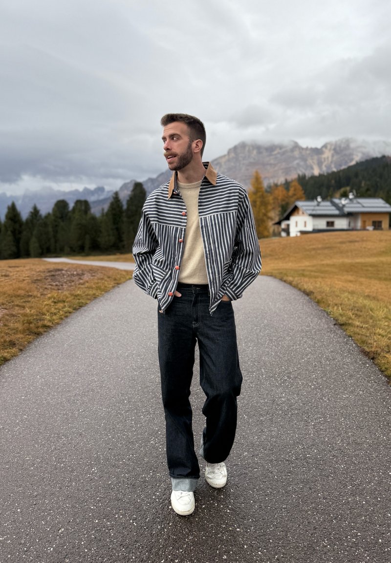 Giacca a righe con colletto color cammello, maglione beige, jeans scuri e scarpe da ginnastica bianche. Ambientazione esterna con montagne e alberi sullo sfondo.