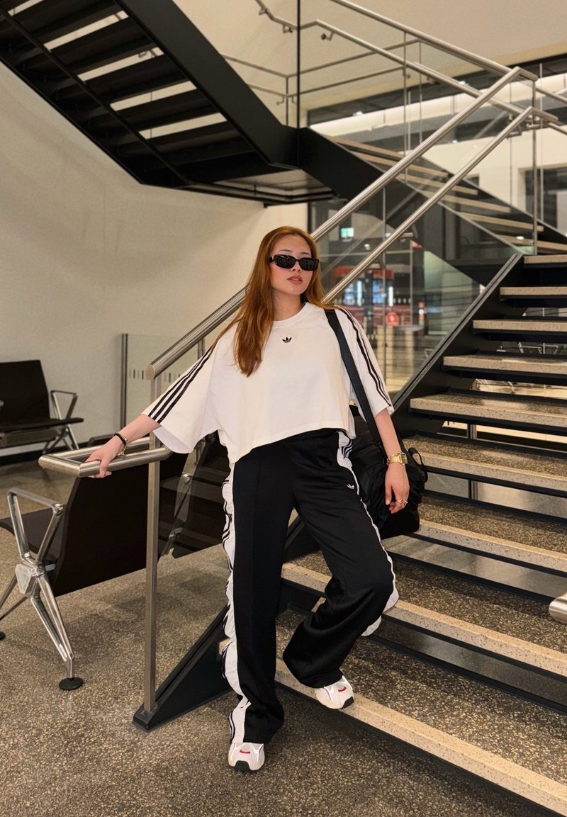 Jeune femme en survêtement Adidas noir et blanc et lunettes de soleil, appuyée sur la rampe d'escalier à l'intérieur d'un bâtiment moderne.
