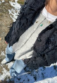 Schwarze, strukturierte Jacke mit ausgefransten Kanten, hellgraues Hemd mit Knöpfen und lockere hellblaue Jeans. Detaillierte goldene Halskette mit sichtbaren Anhängern.