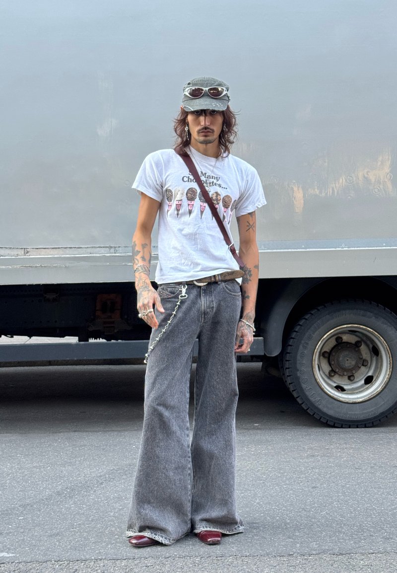 Man with tattoos wearing a green cap with sunglasses, white graphic t-shirt, wide gray jeans, and red shoes standing by a truck.