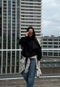 Veste noire oversize avec un col haut, chemise à carreaux légère nouée à la taille, et jeans bleus. L'arrière-plan présente des bâtiments modernes.