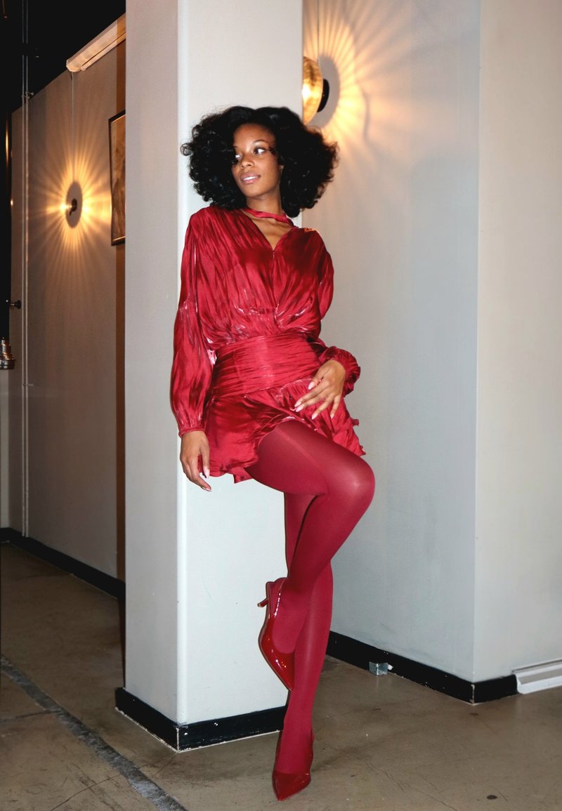 Kvinna med lockigt hår som bär en glänsande röd klänning, matchande röda strumpbyxor och klackar, lutande mot en vit pelare i en svagt upplyst korridor.