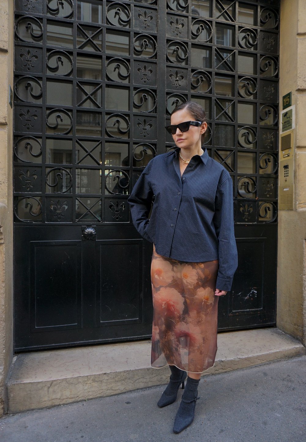 Camisa de gran tamaño a rayas blanco y negro, falda sheer con estampado floral, botas de tobillo grises, gafas de sol, de pie contra una puerta de hierro forjado.