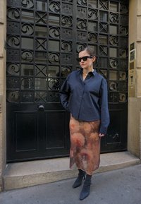 Camisa de gran tamaño a rayas blanco y negro, falda sheer con estampado floral, botas de tobillo grises, gafas de sol, de pie contra una puerta de hierro forjado.