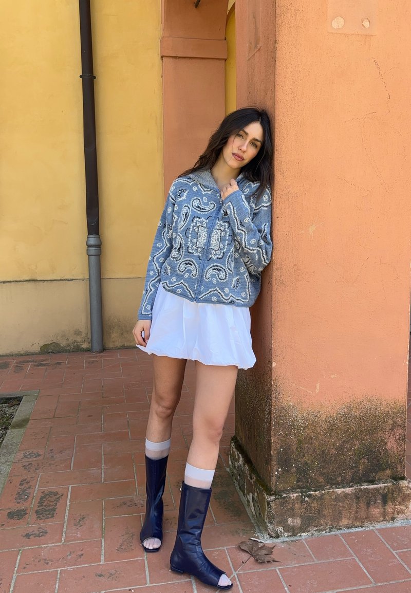 Femme aux longs cheveux bruns appuyée contre un mur orange, portant un pull bleu à motifs, une jupe blanche, des chaussettes transparentes et des bottines ouvertes bleu marine sur un sol carrelé.