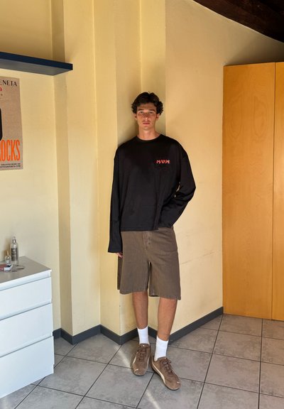 Camisa de manga larga negra con logo rojo, pantalones cortos de pana marrón, calcetines blancos y zapatillas marrones. Pies planos, de pie en una habitación con paredes claras.