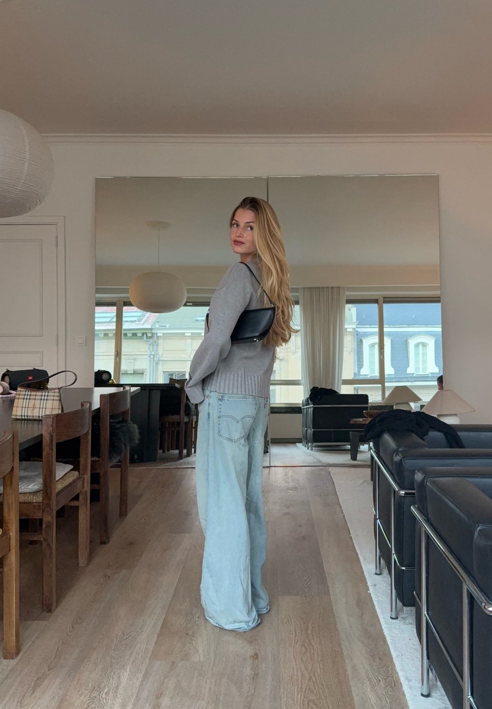 Un pull gris clair et un jean large bleu clair ; un sac à main noir porté sur une épaule. Des surfaces réfléchissantes et des meubles modernes en arrière-plan.