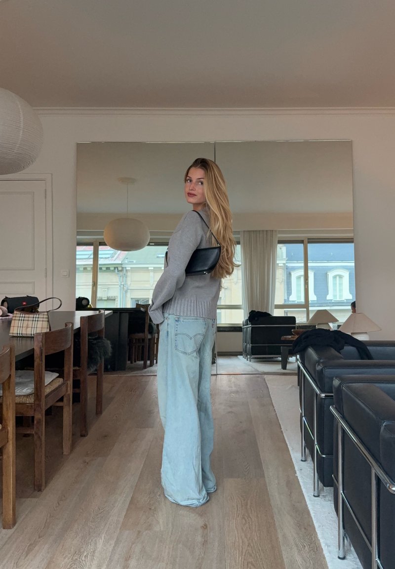 Lichtgrijze trui en lichtblauwe wijde jeans; een zwarte handtas wordt op één schouder gedragen. Reflecterende oppervlakken en moderne meubels op de achtergrond.