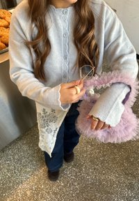 Cardigan grigio chiaro con bordi smerlati, pantaloni di denim blu e una borsa lavanda con volant. Tiene degli occhiali in una mano, mostrando gli anelli sulle dita.