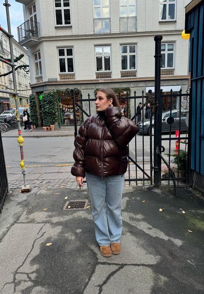 Chaqueta acolchada marrón con un acabado brillante, con cuello alto y un corte oversize. Combinada con vaqueros de pierna ancha de color azul claro y botas marrones.