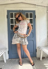 Biela grafická tričko s vintage potlačou, šedá kvetinová mini sukňa a béžová pletená kabelka. Čierne slnečné okuliare a čierne sandále na remienky.