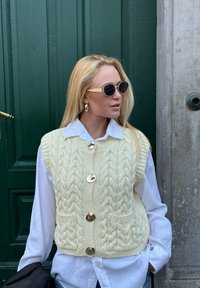 Sweter bez rękawów w kolorze kremowym z wzorem warkoczowym, z ozdobnymi złotymi guzikami i kieszeniami, nałożony na białą koszulę; dopełniony okrągłymi czarnymi okularami przeciwsłonecznymi.