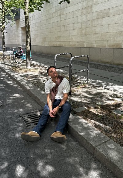 Joven sentado en un bordillo de concreto, vistiendo una camisa blanca de manga corta y jeans azules, con un suéter marrón colgado sobre los hombros.