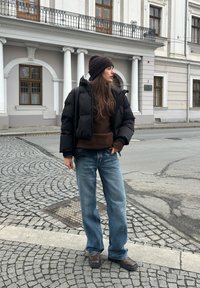 Doudoune noire, pull marron, jean bleu ample et bottes marron. Debout sur une rue pavée avec un bâtiment historique en arrière-plan.