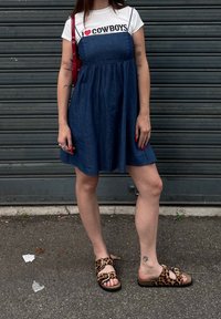 Denimová šaty s nariasenou sukňou, prevrstvené cez biele tričko s čiernym a červeným textom. Hnedé sandále s leopardím vzorom a prackovými detailami.