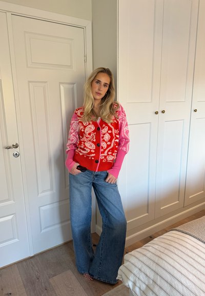 Cárdigan con estampado paisley rojo y rosa, con botones delanteros, combinado con vaqueros de pierna ancha en azul claro. Pared blanca y armario en el fondo.