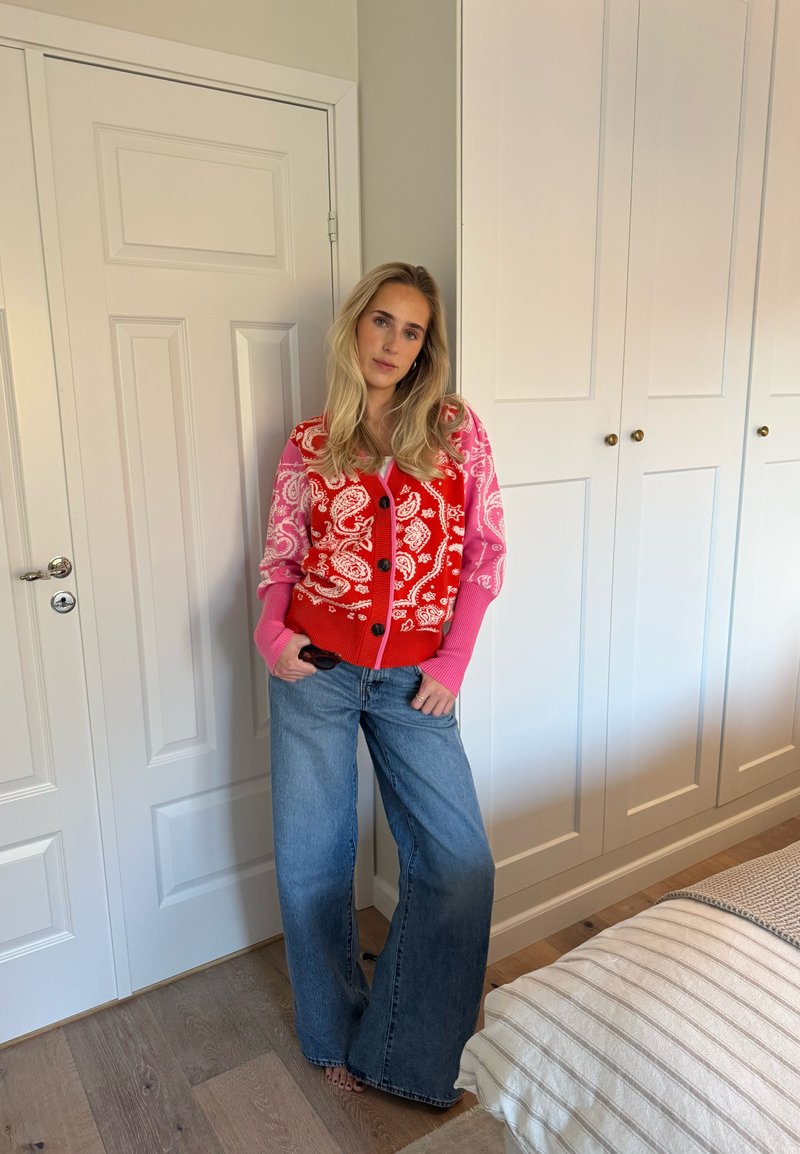 Cardigan à motifs paisley rouges et roses, fermé par des boutons sur le devant, associé à un jean large bleu clair. Mur blanc et armoire en arrière-plan.