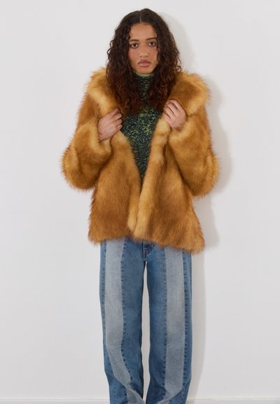 Abrigo de piel sintética en marrón dorado con mangas anchas y un corte relajado, usado sobre un cuello de tortuga verde con estampado y combinado con jeans a rayas.