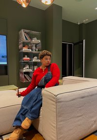 Κόκκινο πλεκτό πουλόβερ και τζιν καλύπτες, καθισμένο σε ένα ανοιχτόχρωμο καναπέ σε έναν πράσινο χώρο. Το υπόβαθρο περιλαμβάνει μια μονάδα ραφιού και έργα τέχνης.