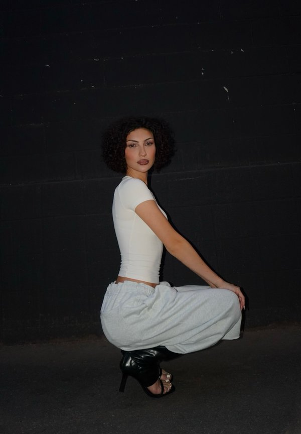 Donna con capelli ricci che indossa una canottiera bianca, pantaloni grigi e scarpe nere con tacchi alti accovacciata contro una parete scura.
