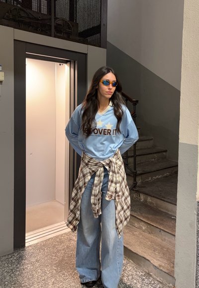 Mujer con cabello largo y oscuro, usando gafas de sol, una camisa azul con texto, una camisa de cuadros atada y jeans anchos, de pie cerca de un ascensor abierto y escaleras.