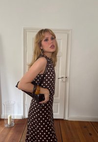 Bruine jurk met stippen, een aansluitende silhouet, mouwloos ontwerp en een soepele textuur. Gecombineerd met een kleine zwarte tas en een amberkleurige armband.