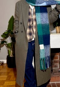 Longue veste vert olive portée sur une chemise à carreaux, un jean denim bleu foncé et une écharpe à carreaux bleu multiton avec des franges. En arrière-plan, des plantes et une cheminée.