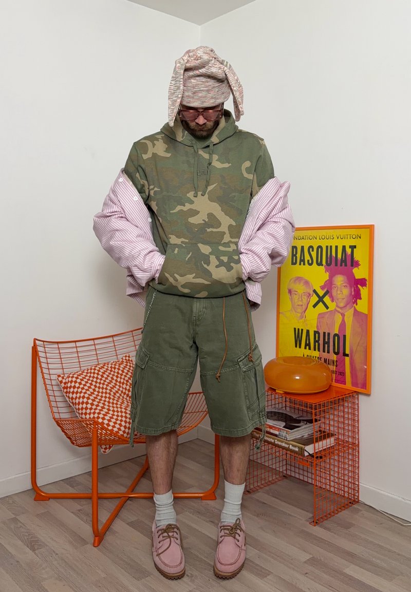 Hombre con sudadera con capucha de camuflaje, pantalones cortos cargo verdes, zapatos rosas, chaqueta rosa caída de los hombros y gorro de punto rosa con orejas caídas, de pie cerca de una silla naranja.