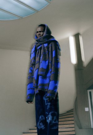 Un pulover supradimensionat, bleu și negru, cu detalii din franjuri, asortat cu pantaloni texturați închisi la culoare, cu modele grafice, într-un interior minimalist.