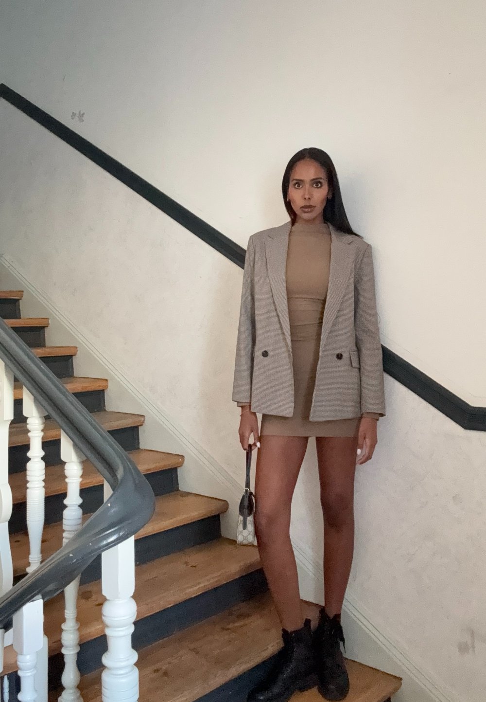 Blazer em padrão pied-de-poule cinza sobre um vestido justo bege, combinado com botas de tornozelo pretas. A modelo segura uma bolsa pequena, de pé em escadas de madeira.