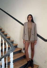 Blazer em padrão pied-de-poule cinza sobre um vestido justo bege, combinado com botas de tornozelo pretas. A modelo segura uma bolsa pequena, de pé em escadas de madeira.