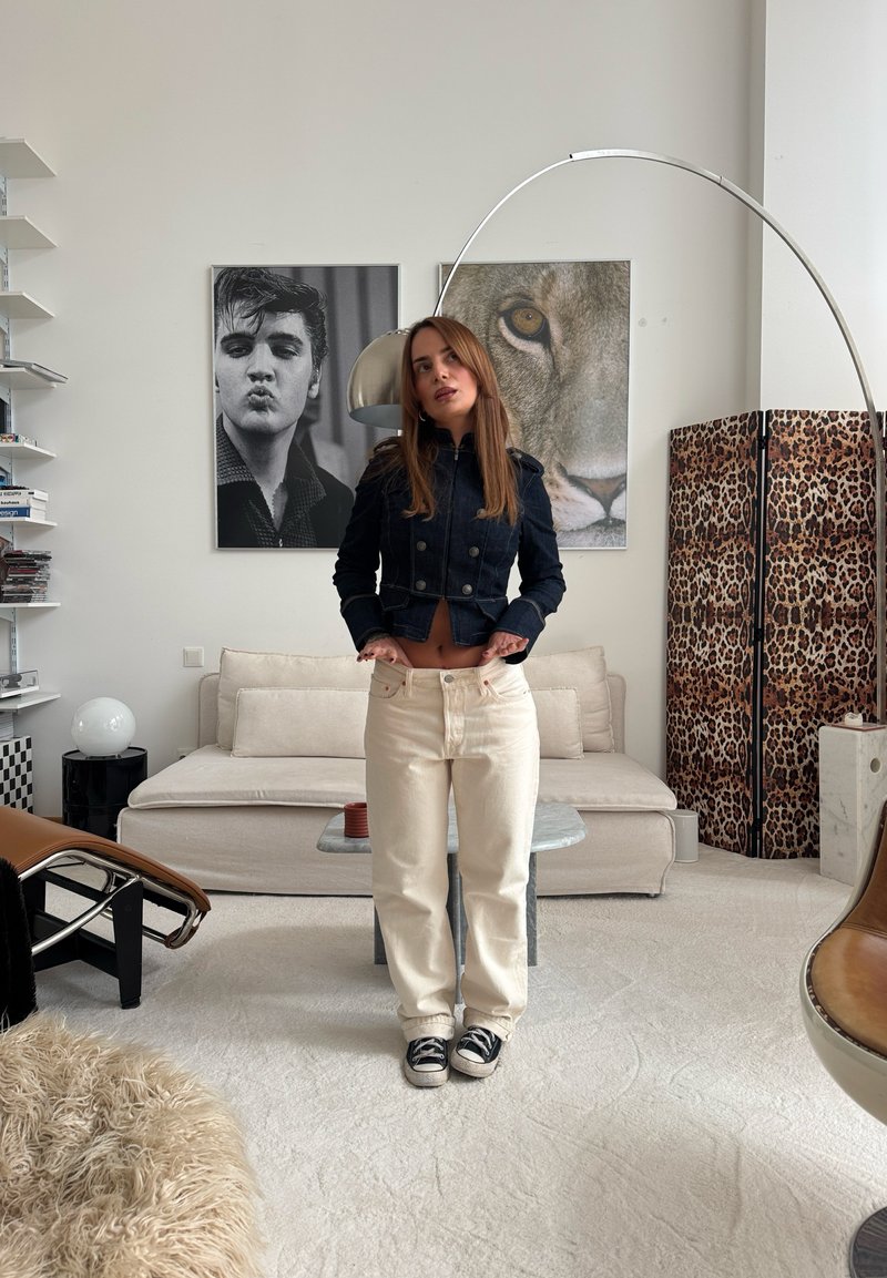 Femme en vêtements décontractés debout dans un salon moderne avec un canapé beige, un portrait en noir et blanc, une photo de lion et un paravent à motif léopard.