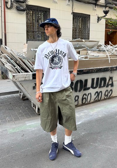 Camiseta blanca con gráfico negro de "New York", pantalones cortos verde oliva, calcetines blancos, zapatillas azules, de pie frente a un fondo de escombros de construcción.