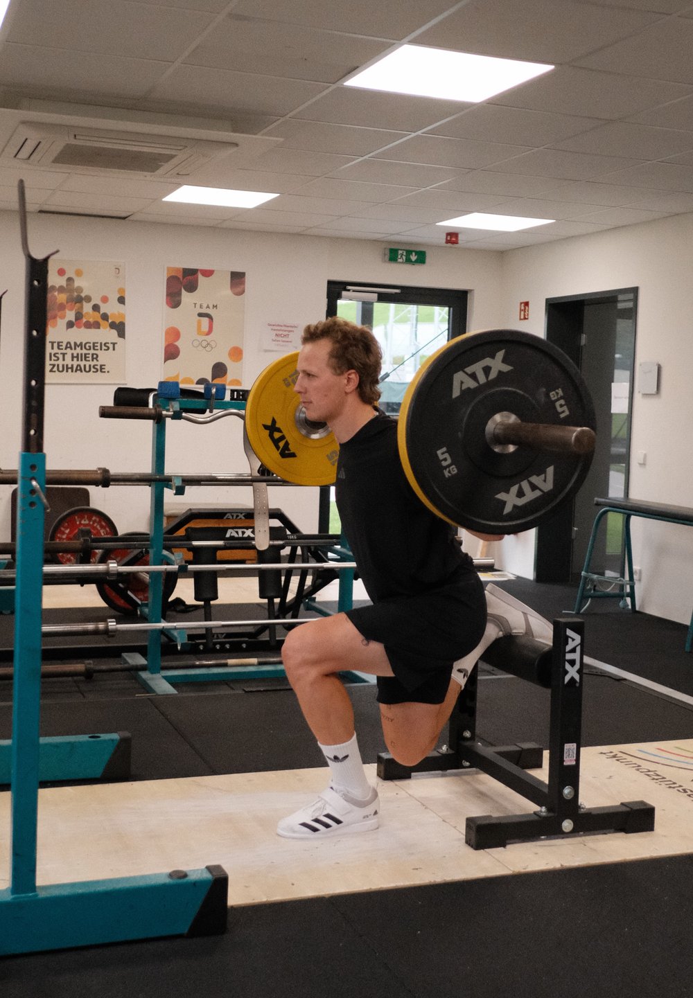Vīrietis veic svaru Bulgāru šķeltās squats ar stieni, uz kura ir dzeltenas un melnas svaru plāksnes, sporta zālē.