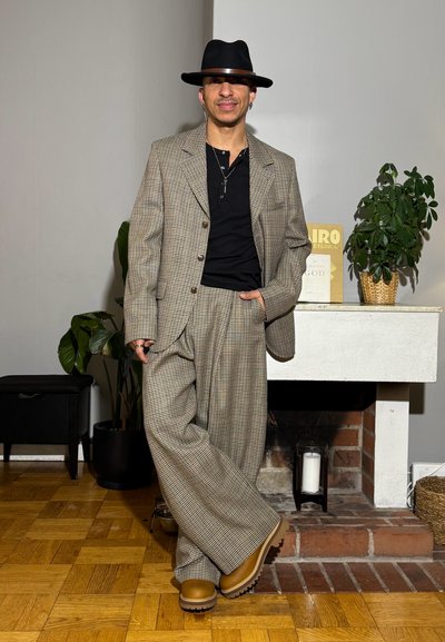 Hombre con traje de cuadros, camisa negra y sombrero negro apoyado en una chimenea blanca con planta verde y libros en la repisa.