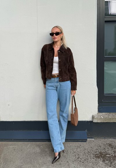 Mujer con chaqueta marrón oscuro, top blanco, vaqueros azul claro, tacones negros, gafas de sol negras, pendientes dorados y sujetando bolso marrón, está apoyada contra una pared.