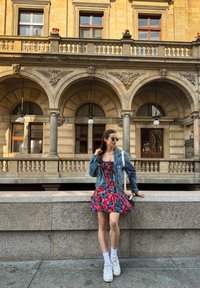 Φορεμάτι με λουλουδάτο σχέδιο σε κόκκινες και μπλε αποχρώσεις, τζιν μπουφάν, λευκά αθλητικά παπούτσια; στέκεται μπροστά σε μια πέτρινη αψίδα με την καμπυλωτή πρόσοψη ενός κτηρίου στο φόντο.