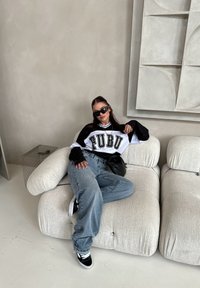 Μαύρη και λευκή φούτερ μπλούζα FUBU, χαλαρές μπλε τζιν παντελόνες, μαύρα γυαλιά ηλίου, καθισμένος σε μια αφράτη ανοιχτόχρωμη καναπέ σε έναν μινιμαλιστικό χώρο.