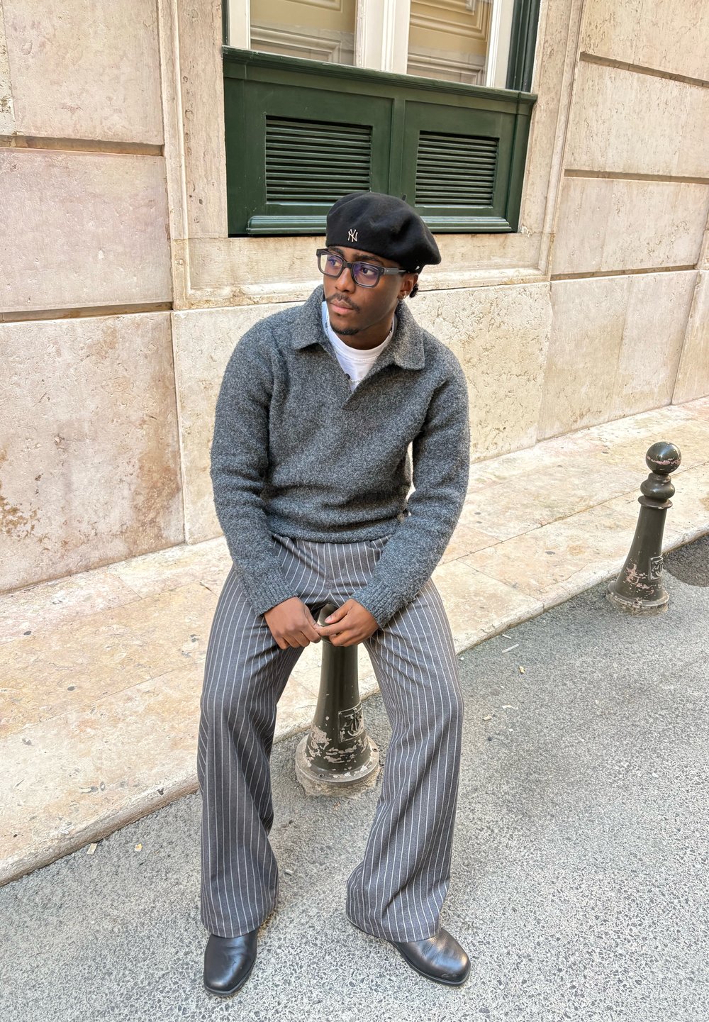 Pull en laine gris avec un col, pantalon gris à larges rayures, béret noir, bottes en cuir noir, assis sur un poteau en métal.