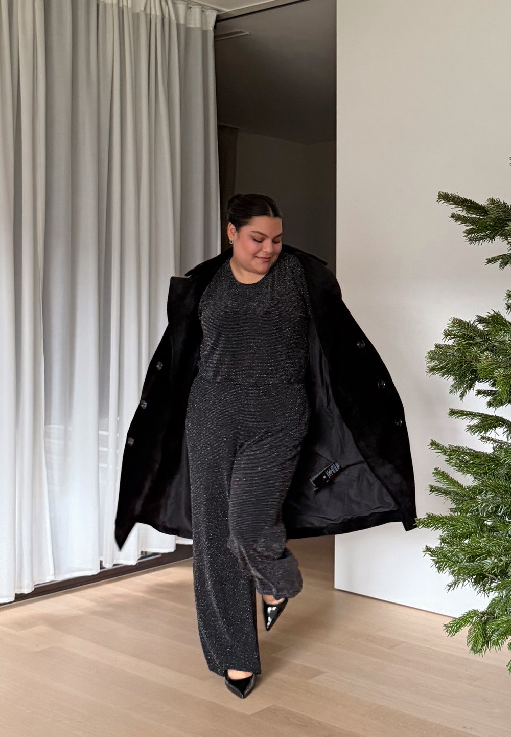 Sort, tekstureret jumpsuit med glitrende detaljer, lagdelt under en oversized sort frakke; modellen har sorte høje hæle med spids tå. Indendørs omgivelser med grønne planter.