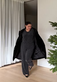 Sort, tekstureret jumpsuit med glitrende detaljer, lagdelt under en oversized sort frakke; modellen har sorte høje hæle med spids tå. Indendørs omgivelser med grønne planter.