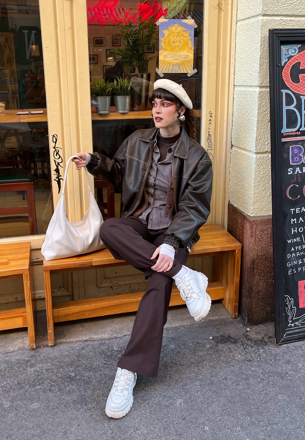 Mannequin portant une veste en cuir marron sur une chemise rayée, un pantalon marron et des baskets blanches épaisses. Tenant un sac crème, avec un béret.